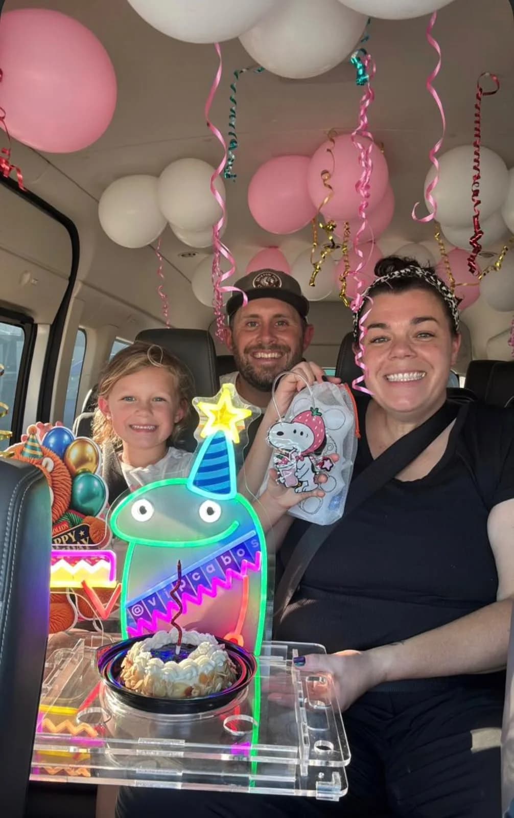 A family smiling inside a VIP Transportation van decorated with pink and white balloons, holding a birthday cake and a glowing sign