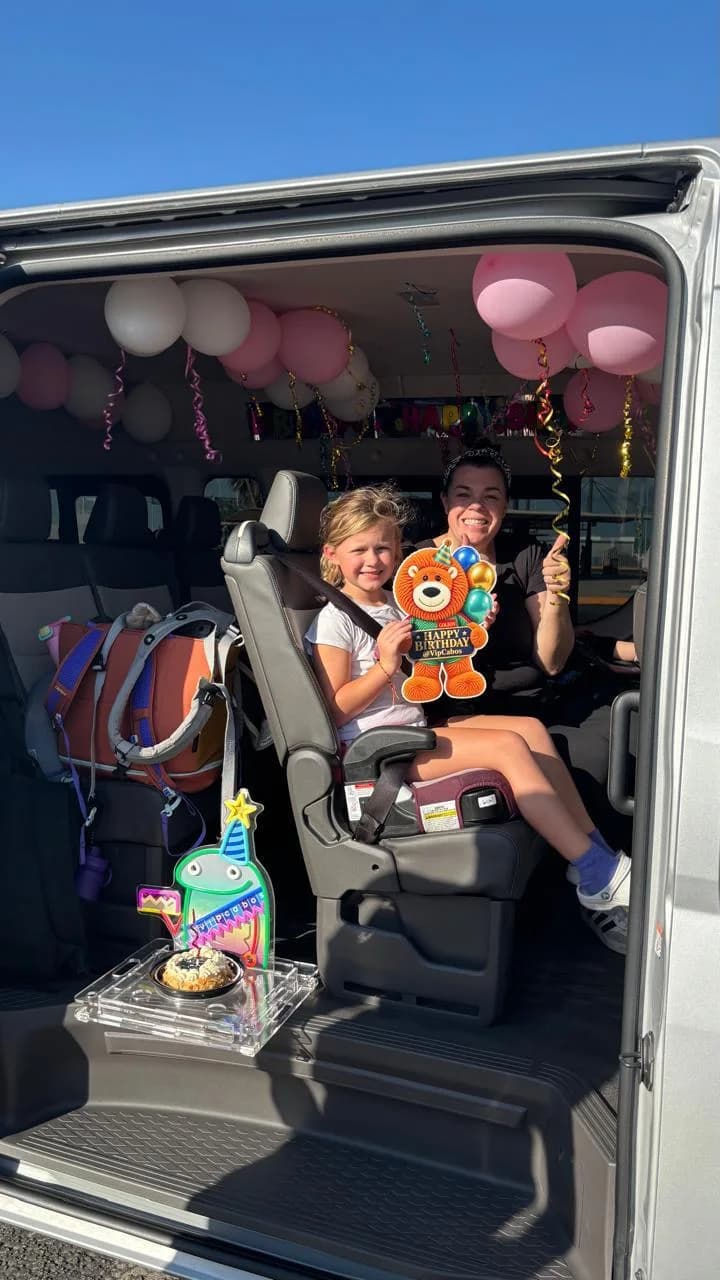A smiling girl in her booster seat holding a Goleón birthday sign as the van door opens